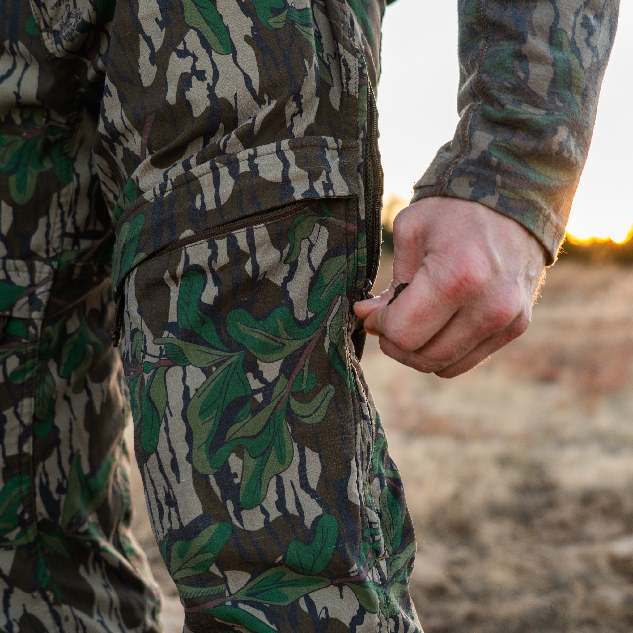 Hunter adjusting side vent zipper on North Mountain Gear Advantis lightweight Mossy Oak Greenleaf camo hunting pants during a hunt.